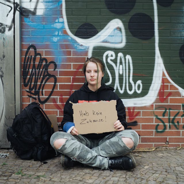 Ein junges obdachloses Mädchen sitzt vor einer Graffiti-Wand und hält ein Pappschild mit der Aufschrift„ Ich habe kein Zuhause“ in der Hand.