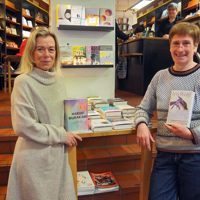 Zwei Frauen stehen in einer Buchhandlung. Jede hat ein Buch in der Hand.