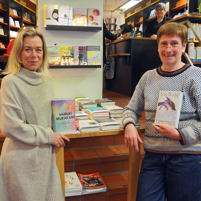 Zwei Frauen stehen in einer Buchhandlung. Jede hat ein Buch in der Hand.