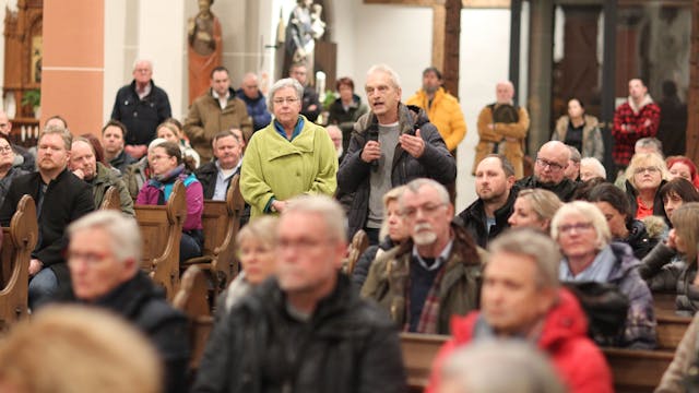 In einer voll besetzen Kirche steht ein Mann und spricht in ein Mikrofon.
