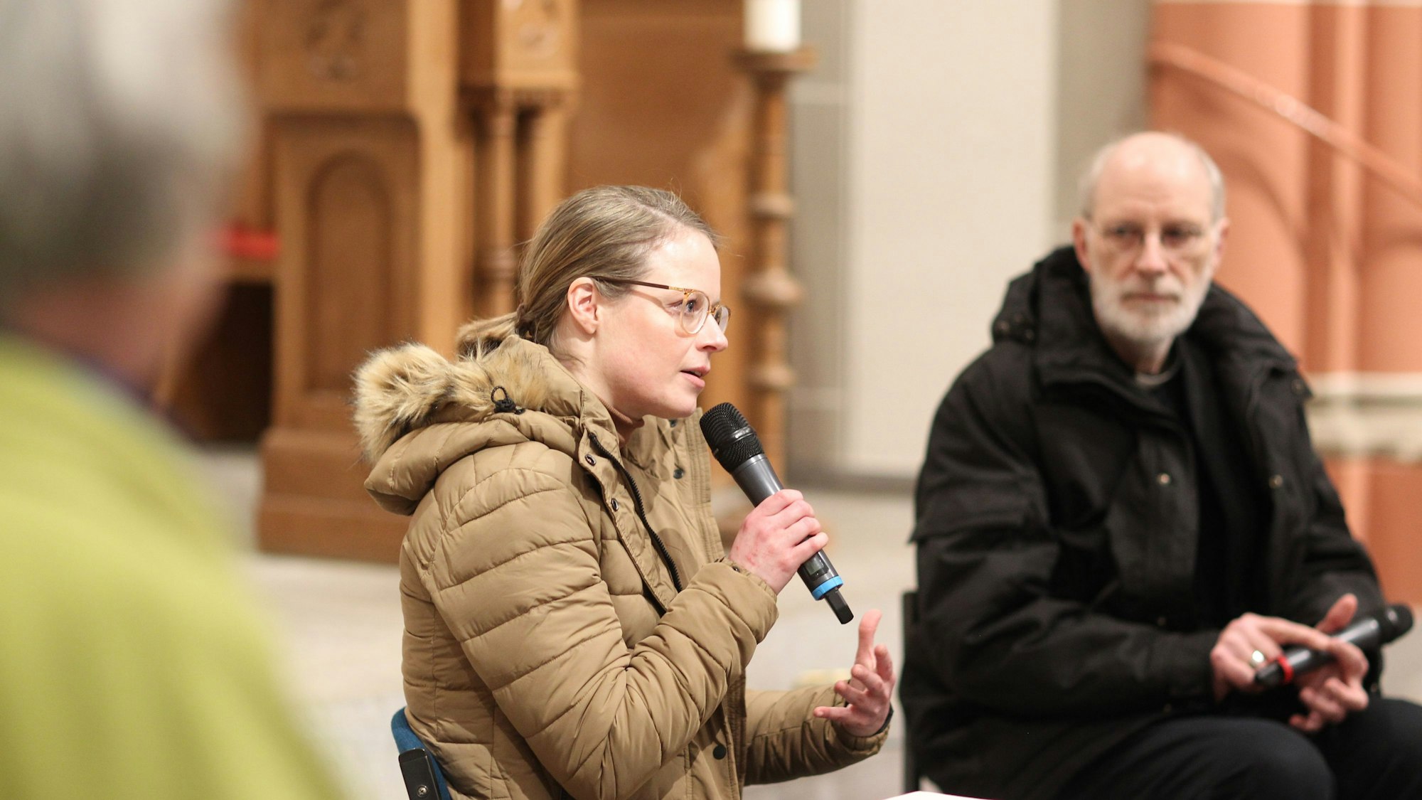 Eine blonde Frau mit Pferdeschwanz und Brille in einem Steppmantel spricht in ein Mikrofon, neben ihr sitzt ein Geistlicher mit grauen Haaren und Bart.