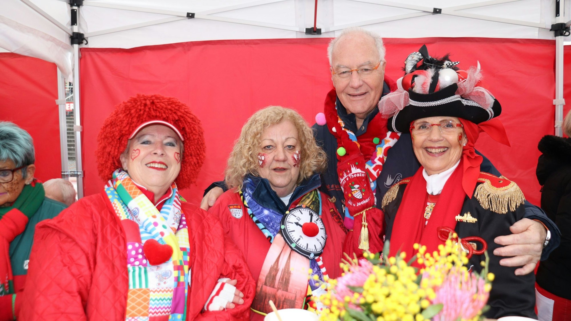 Drei rot-gekleidete Damen und ein Herr mit Karnevalsschal stehen unter einem Pavillon.