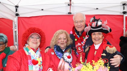 Drei rot-gekleidete Damen und ein Herr mit Karnevalsschal stehen unter einem Pavillon.