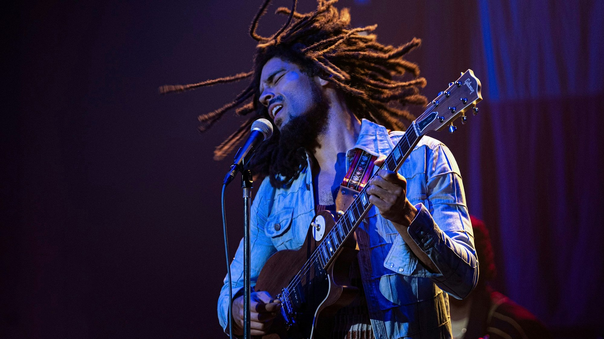 Der britische Schauspieler Kingsley Ben-Adir steht als Bob Marley Gitarre spielend und singend auf der Bühne. Er trägt eine Jeansjacke, seine Rastazöpfe