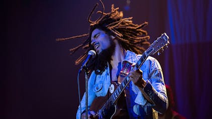 Der britische Schauspieler Kingsley Ben-Adir steht als Bob Marley Gitarre spielend und singend auf der Bühne. Er trägt eine Jeansjacke, seine Rastazöpfe&nbsp;