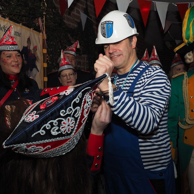 Pulheims Bürgermeister im Blaumann, darunter ein Ringelshirt in den Farben Blau und Weiß. Ein Mitglied der KG Löschgrenadiere versucht, die Handschellen zu lösen.&nbsp;