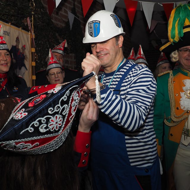 Pulheims Bürgermeister im Blaumann, darunter ein Ringelshirt in den Farben Blau und Weiß. Ein Mitglied der KG Löschgrenadiere versucht, die Handschellen zu lösen.&nbsp;