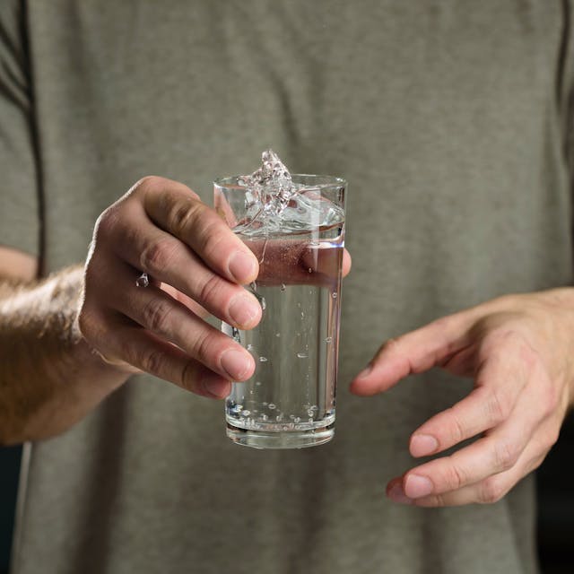 Eine Person hält ein Glas Wasser in der Hand. Das Wasser wirft kleine Wellen, was auf ein Zittern der Person hindeutet.