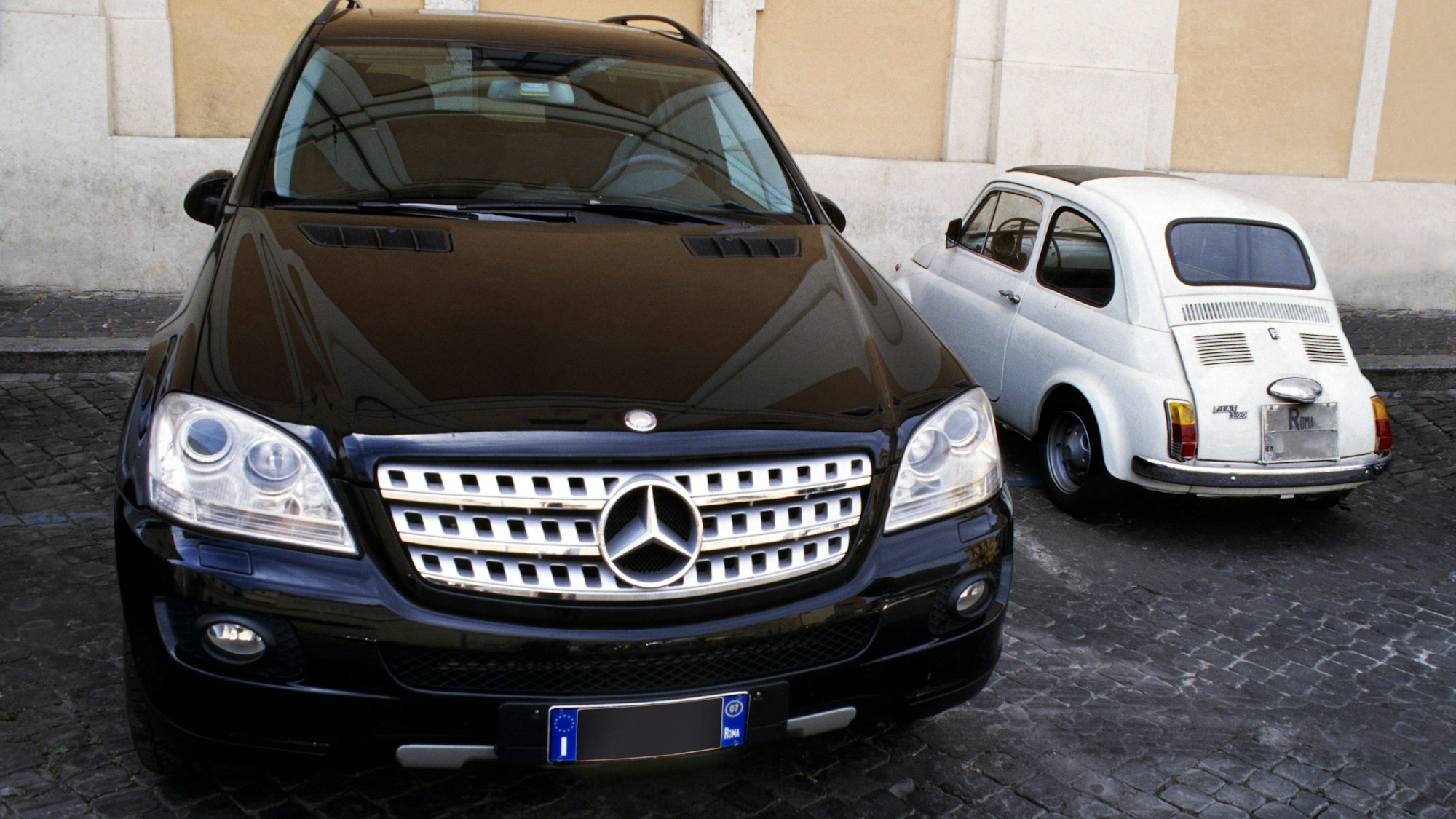 Parkende Autos: Ein großer schwarzer SUV und ein winziger weißer Fiat 500 nebeneinander eingeparkt.