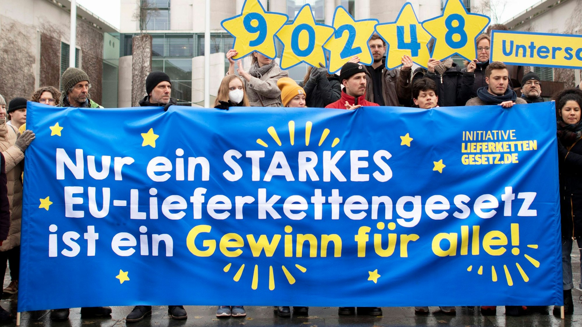 Vor dem Bundeskanzleramt in Berlin hält eine kleine Gruppe von Demonstranten ein blaues Transparent mit der Aufschrift „Nur ein starkes EU-Lieferkettengesetz ist ein Gewinn für alle!“