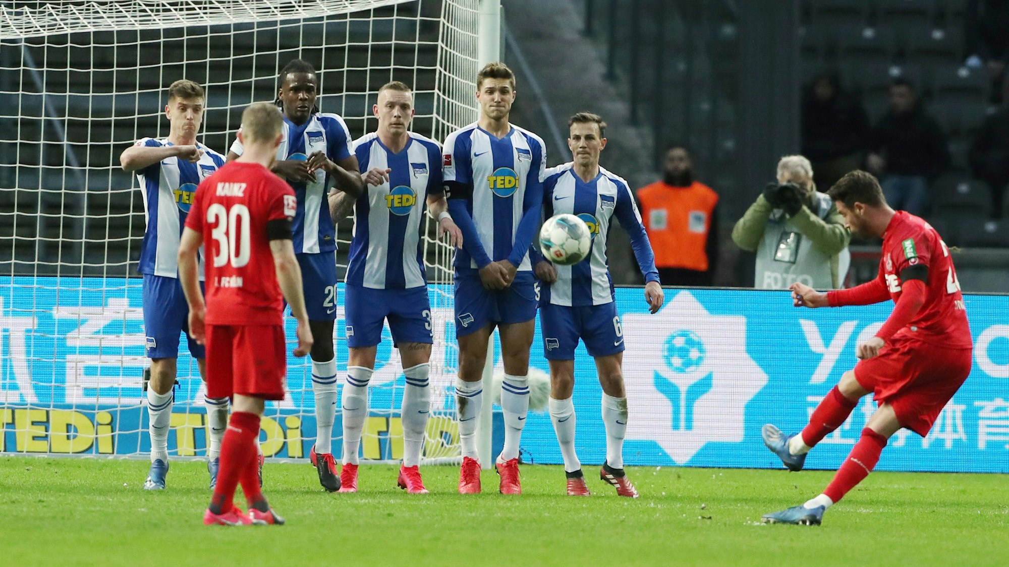 Der ganz in rot gekleidete Profi des 1. FC Köln Mark Uth schießt einen Freistoß über die in Blau-Weiß gekleideten Berliner Spieler, die eine Mauer gebildet haben. Im Hintergrund ist das Tor zu stehen.