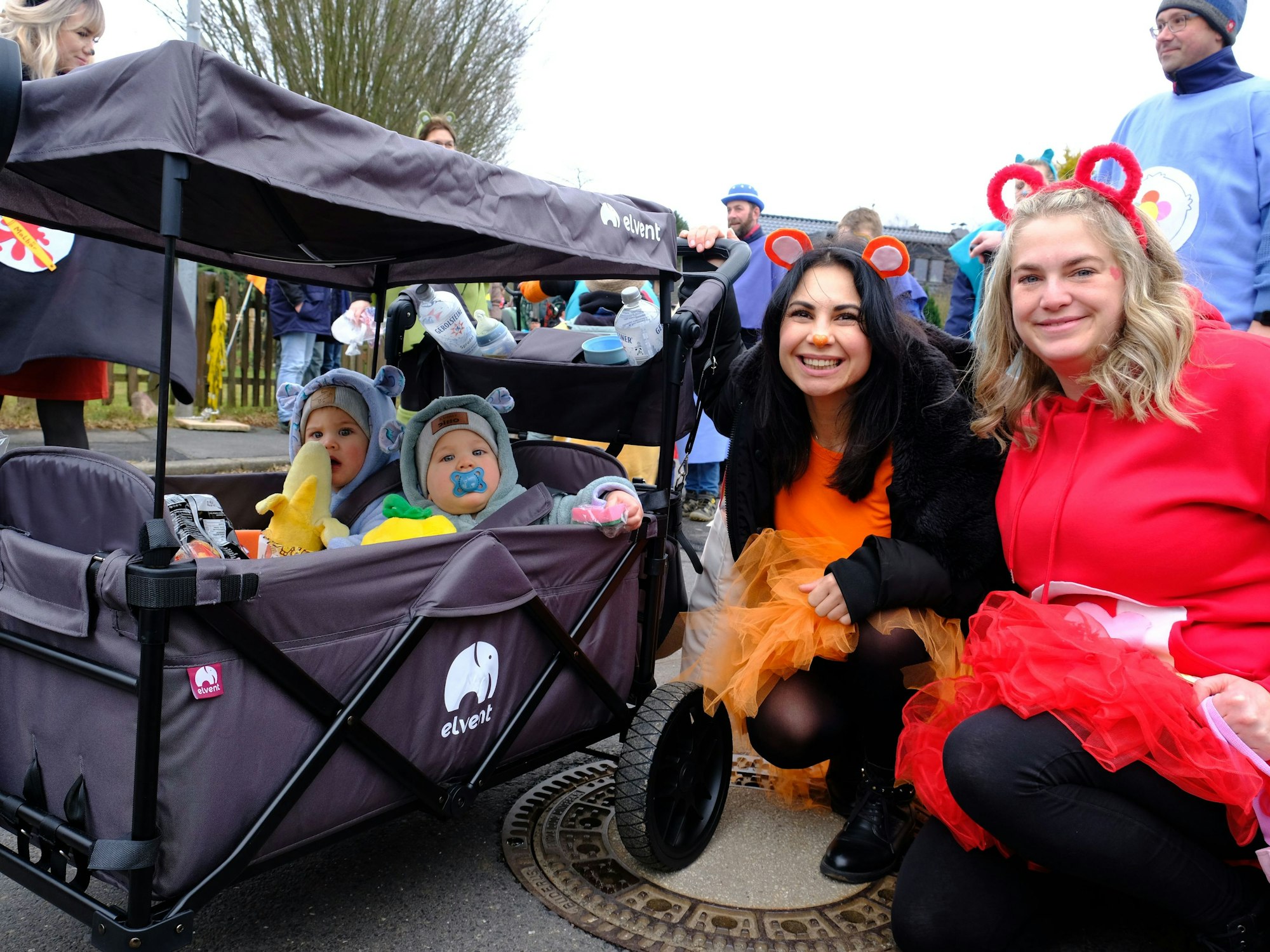 Zwei Kinder sitzen in einem Bollerwagen, zwei Frauen in Kostümen knien daneben.