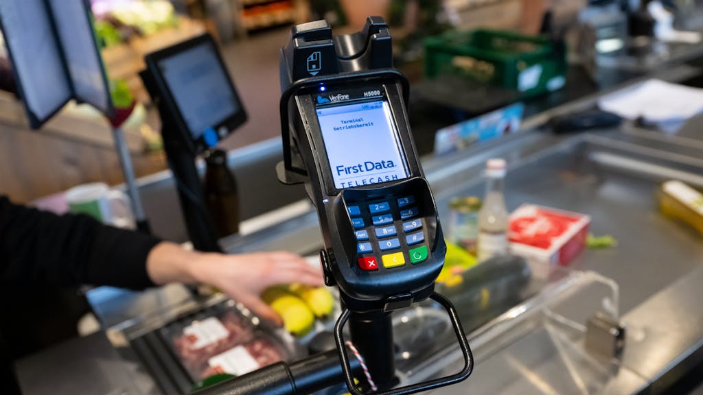 Ein Kassierer scannt in einem Supermarkt an der Kasse die Produkte. Im Vordergrund ist ein EC Terminal zu sehen.