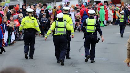 Polizisten gehen auf der Kürtener Bergstraße, auf der der Karnevalszug herannaht. Am Rand stehen kostümierte Jecke.