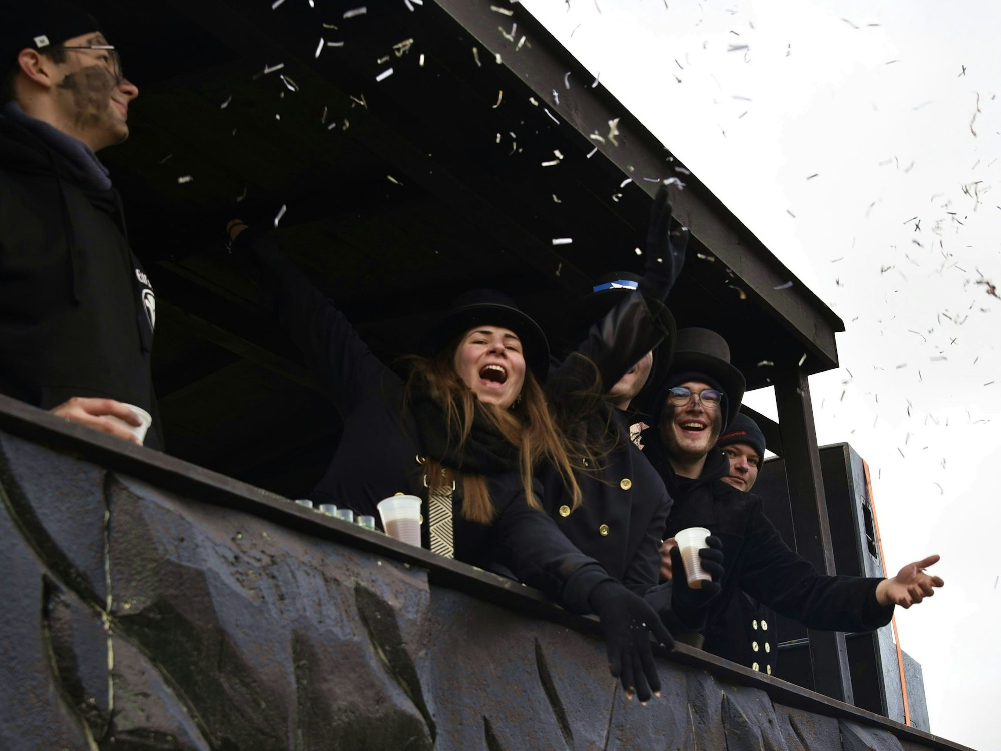 Junge Frauen und Männer feiern und winken von einem schwarzen Karnevalswagen aus.