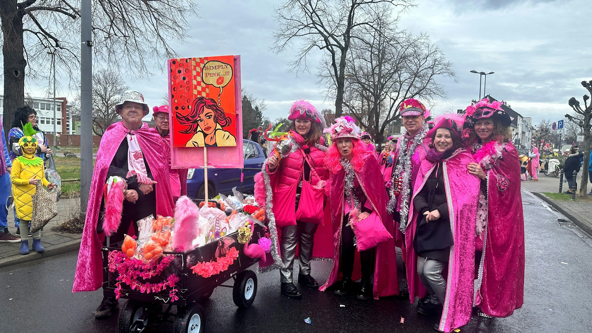 Eine Gruppe pink gekleideter Jecken mit einem Kamellewagen steht auf der Straße.