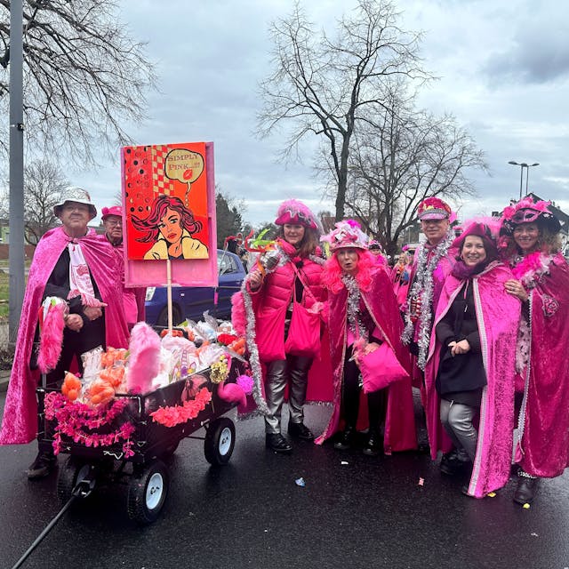 Eine Gruppe pink gekleideter Jecken mit einem Kamellewagen steht auf der Straße.
