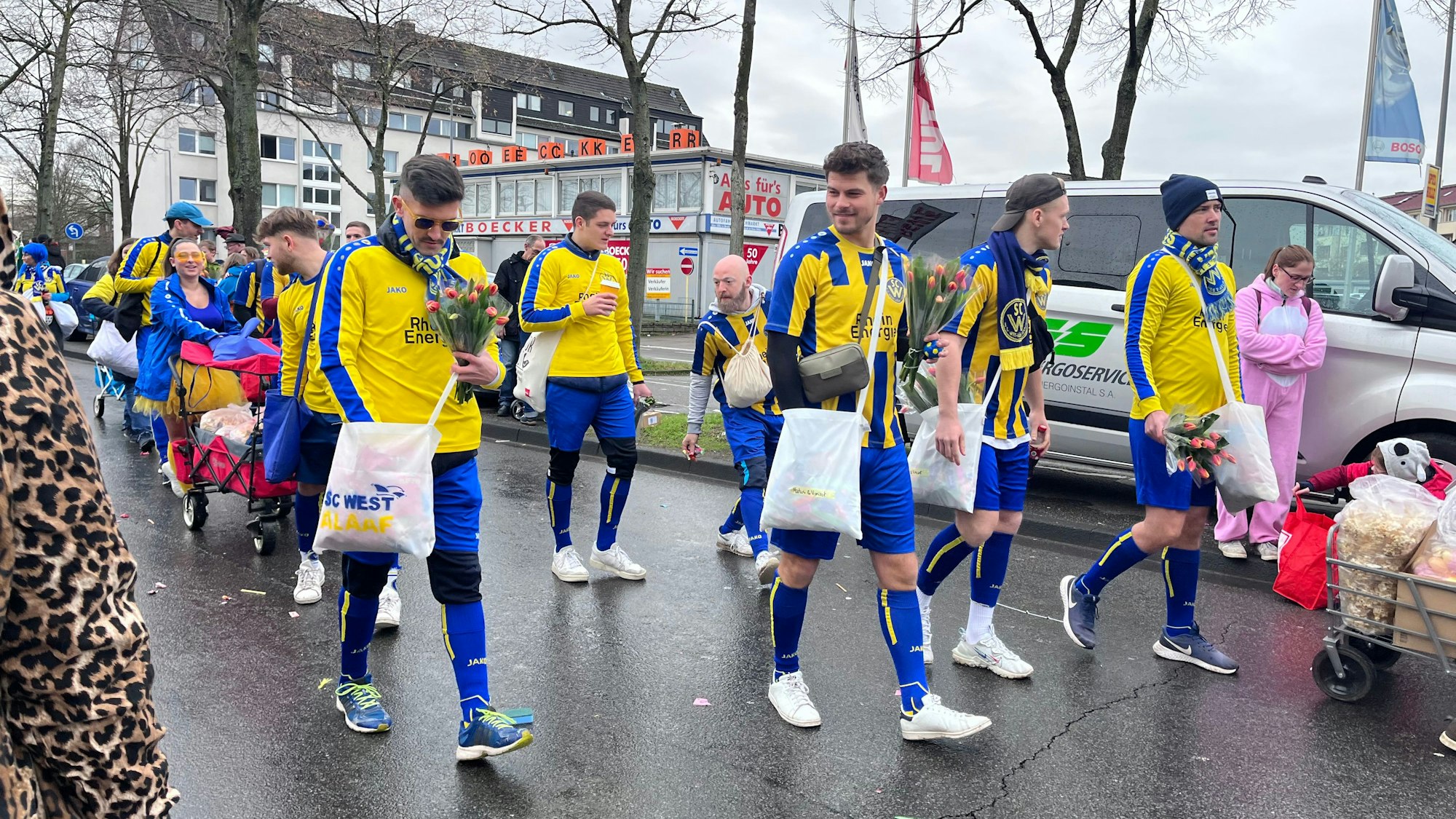 Fußballer in blau-gelben Trikots laufen mit Kamellebüggeln und Strüßjer bewaffnet eine Straße entlang.