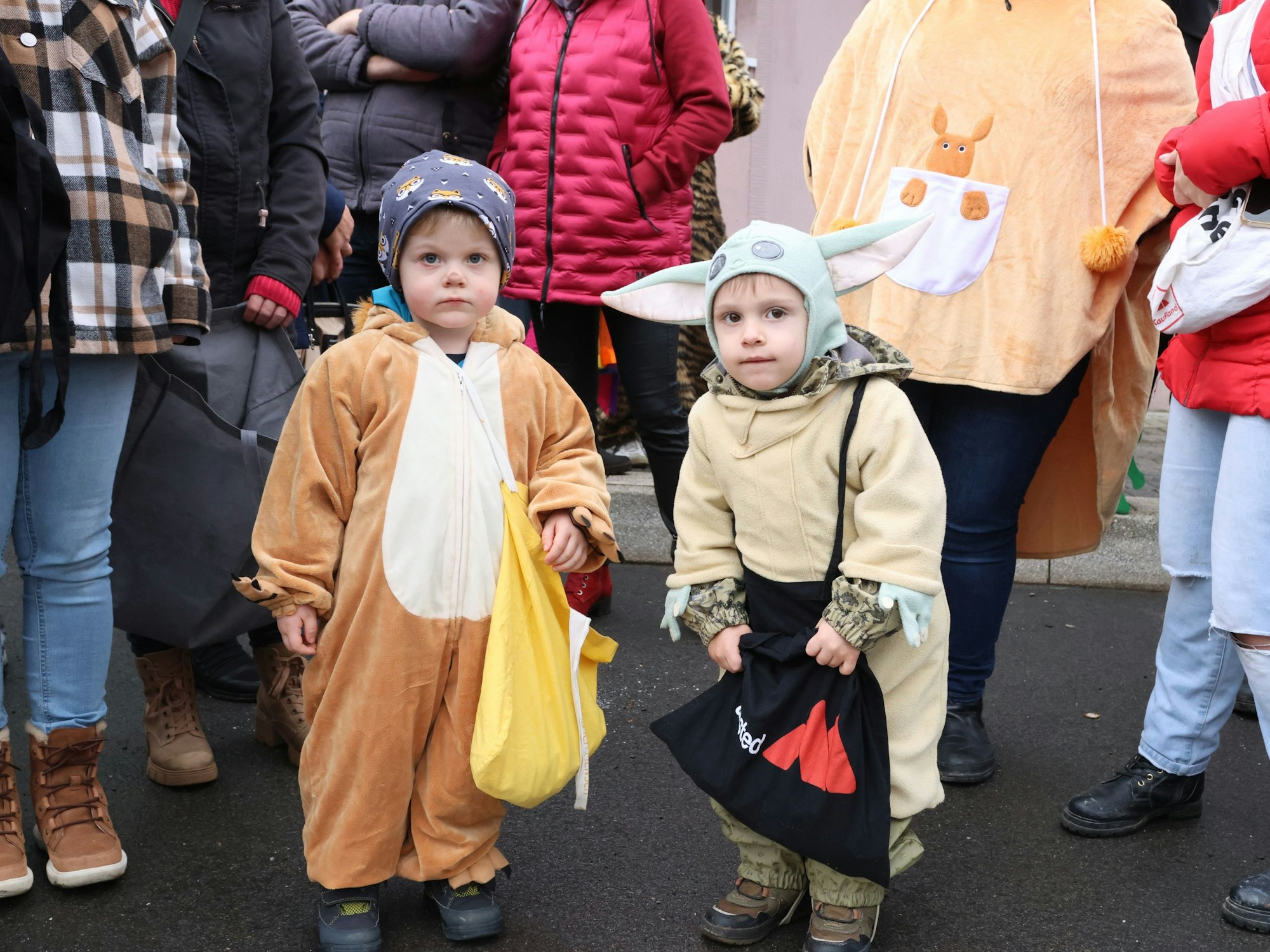 Zwei kleine Kinder in Kostümen stehen mit Beuteln am Rand eines Karnevalsumzuges.