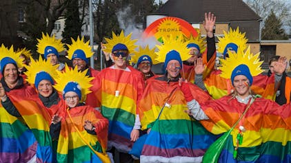 Eine Gruppe von Männern und Frauen mit Regenbogen-Umhängen und Strahlenkronen auf dem Kopf.