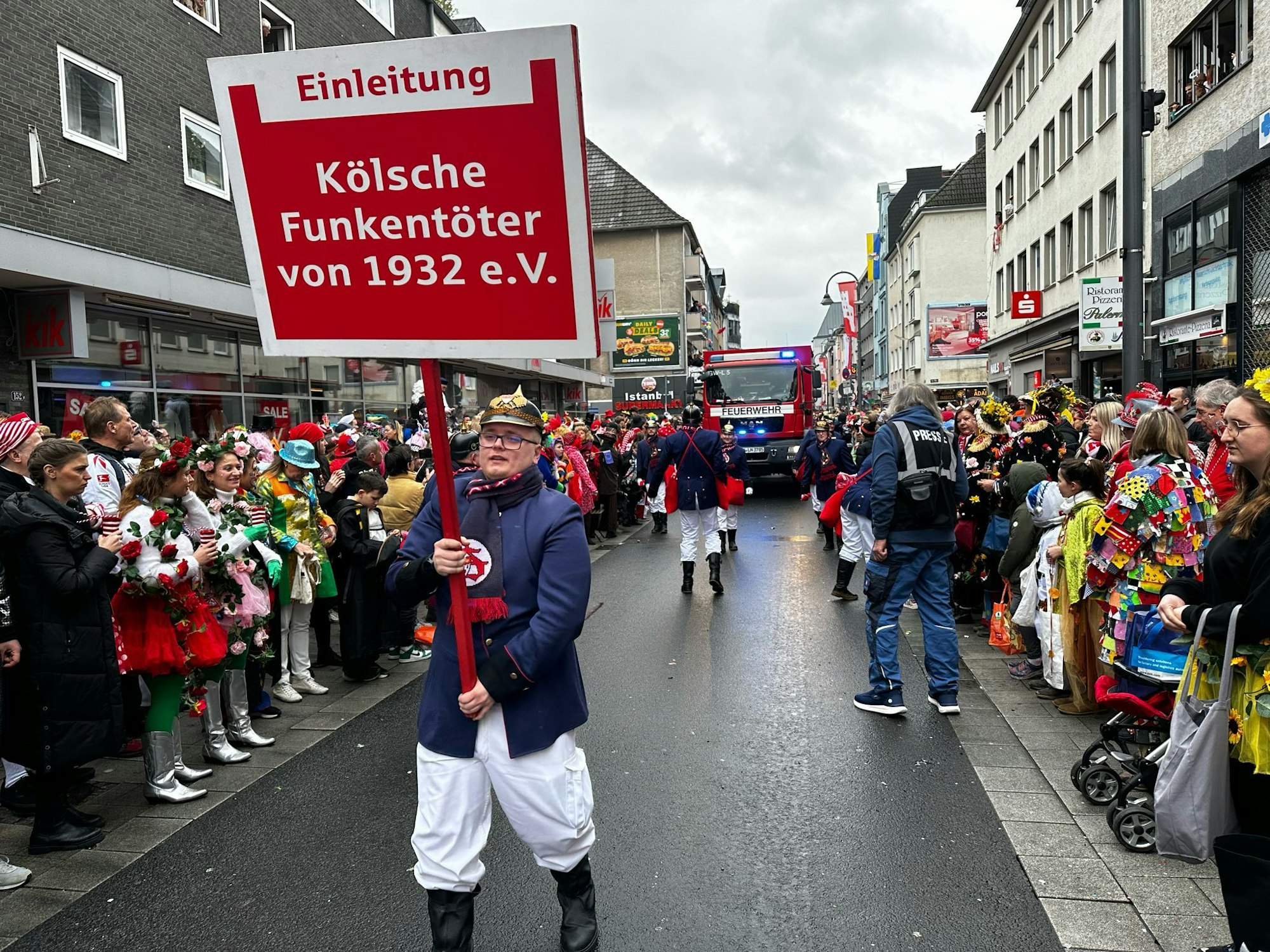 Mann trägt das große Schild einer Kölner Karnevalsgesellschaft.