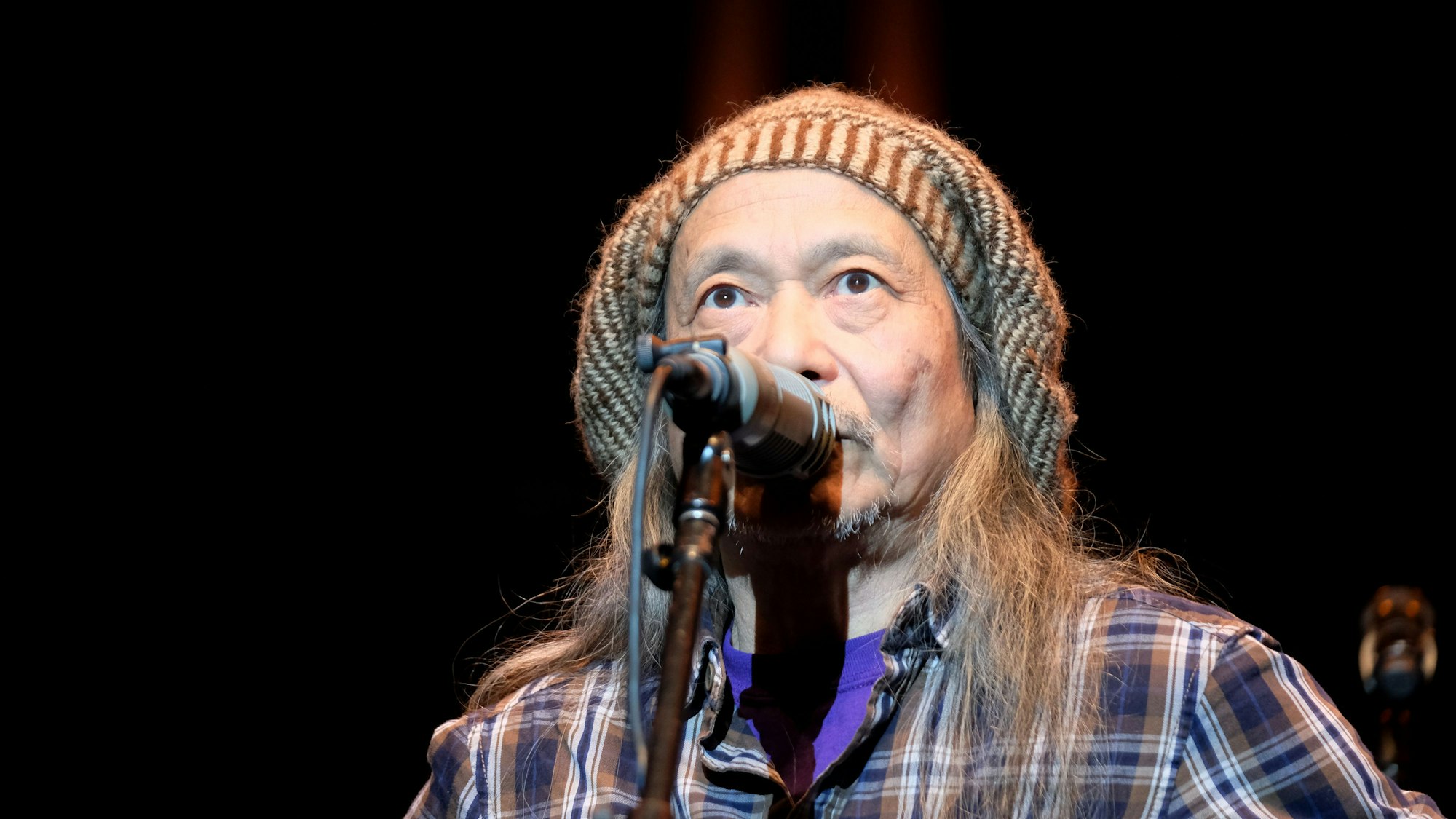 Damo Suzuki steht bei einem Auftritt mit Mütze und länger Mähne auf der Bühne und singt.