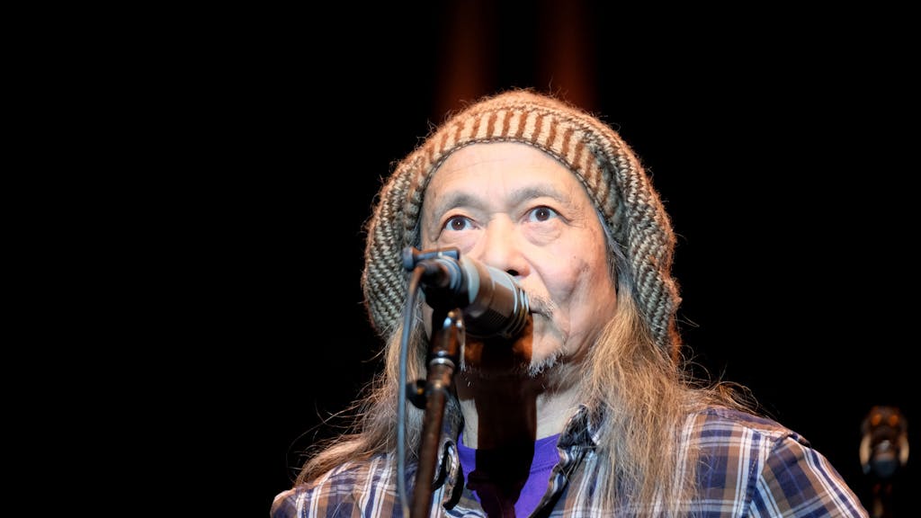Damo Suzuki steht bei einem Auftritt mit Mütze und länger Mähne auf der Bühne und singt.