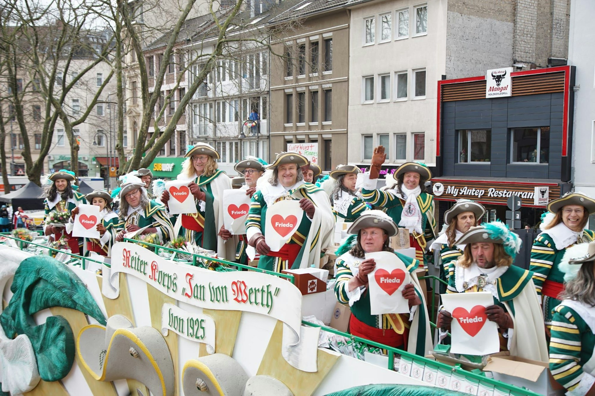 Verkleidete Männer stehen auf dem Karnevalswagen mit dem Schriftzug „Reiter Koprs Jan von Werth“ und halten Herzen mit dem Namen Friede vor sich.