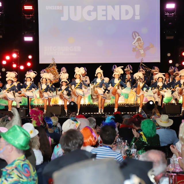 Eine Tanzgarde auf der Bühne eines vollen Saales. Die Besucher sind kostümiert, die Tänzerinnen tragen ihre Gardeuniform.