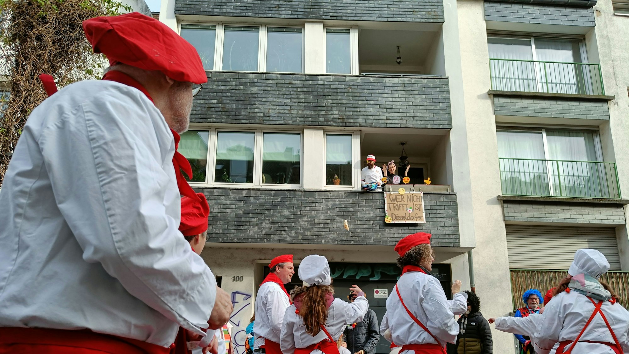 Im Vordergrund ist eine Gruppe in Koch-Kostümen zu sehen. Von einem Balkon hängt ein Pappschild herunter.