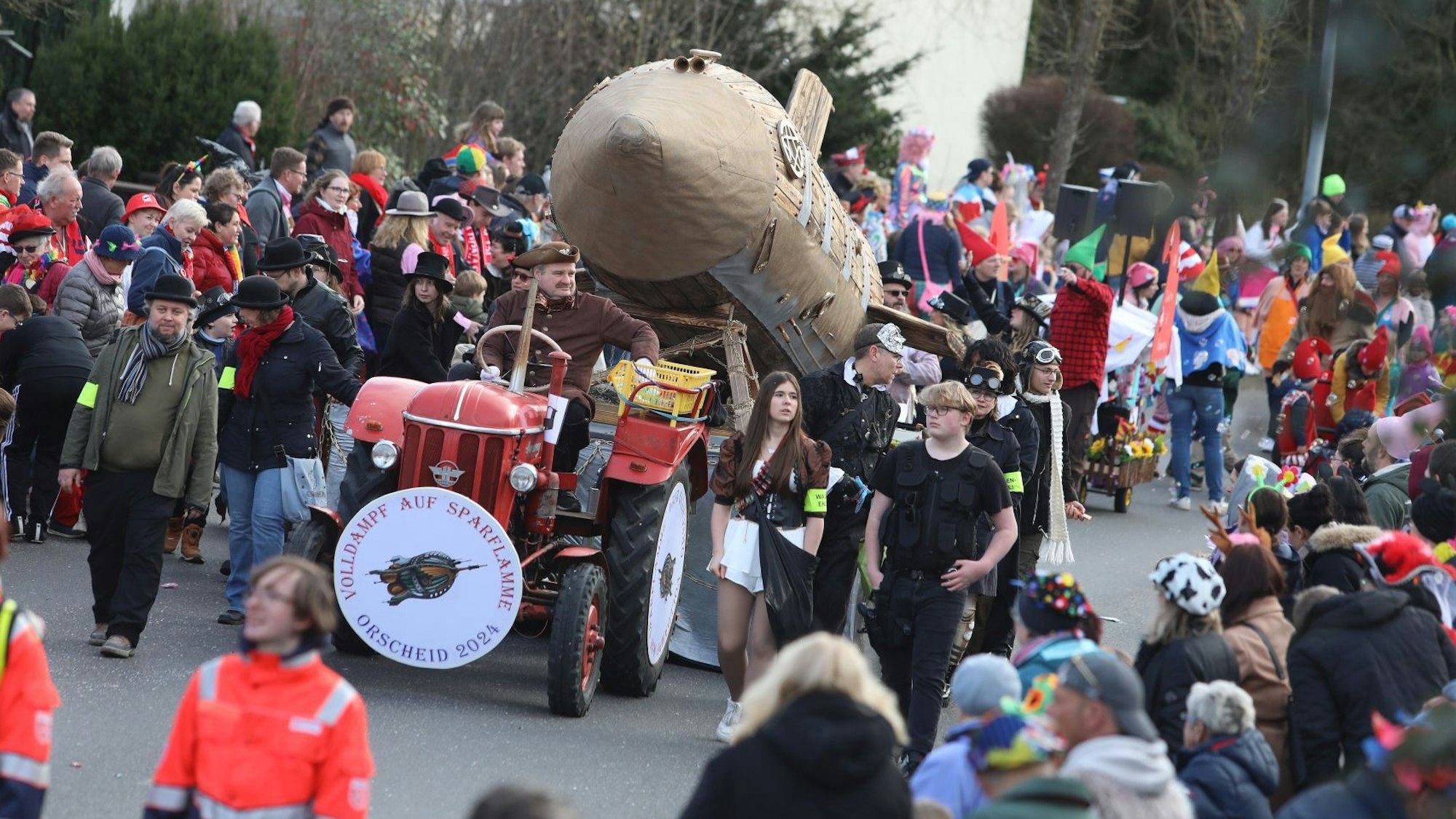 Ein roter Traktor, der eine Holz-Rakete zieht, fährt an vielen kostümierten Zuschauern vorbei.