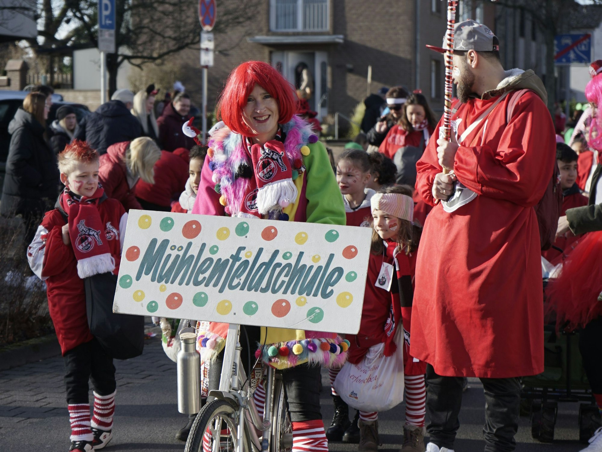 Schulleiterin Ulrike Fuchs mit roter Perücke auf einem Fahrrad und einem Schild mit der Aufschrift Mühlenfeldschule.
