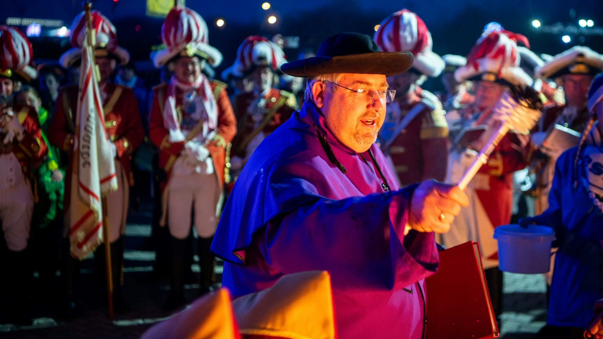 Michael Pesch trägt einen Hut und das violette Gewand eines Pastors und hält eine Predigt. Im Hintergrund stehen die Karnevalisten und hören zu.