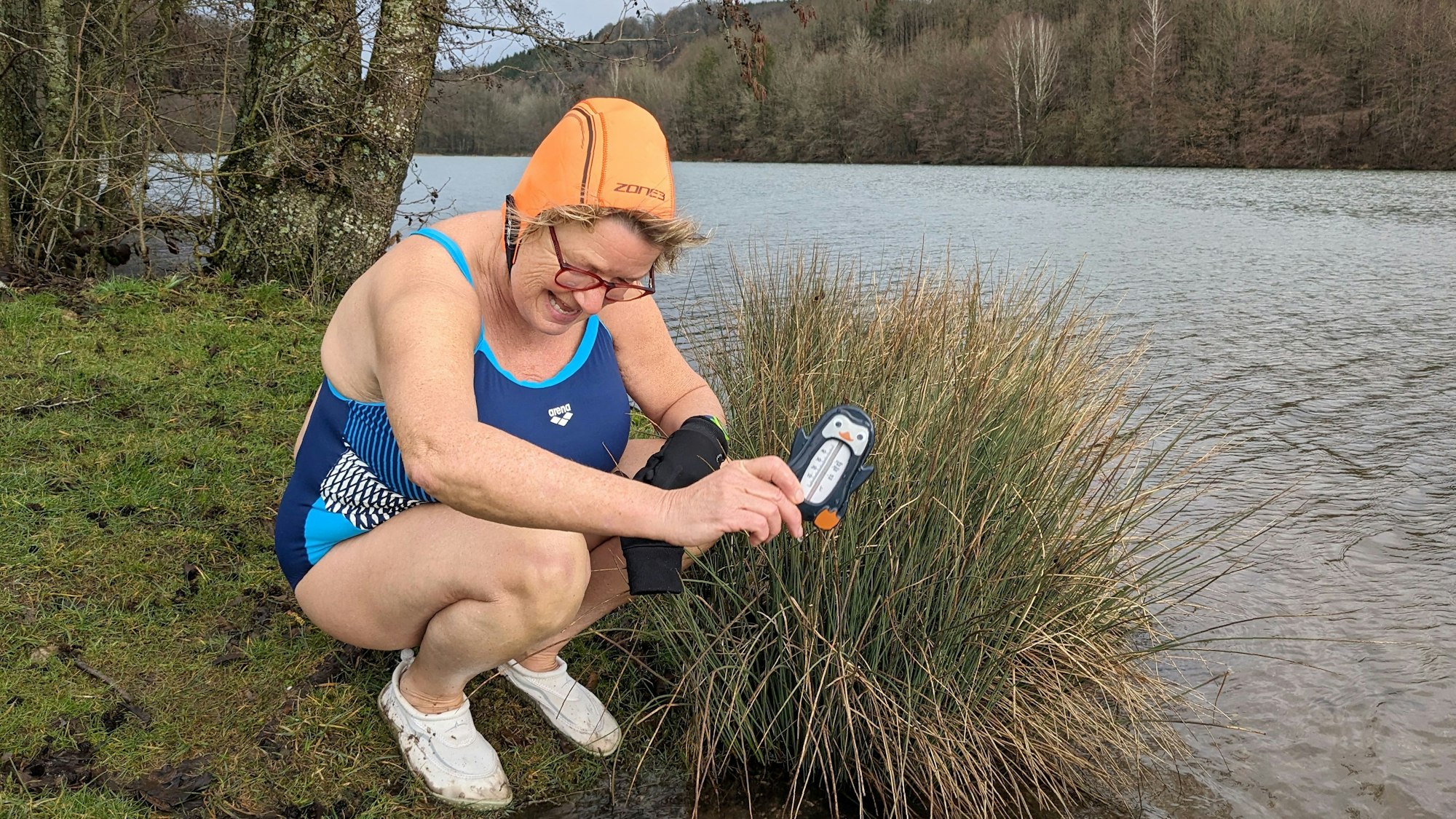 Winterbaderin Jutta Lorenz kniet mit einem Thermometer am Ufer des Freilinger Sees. Sie trägt einen blauen Badeanzug und eine orangefarbene Badehaube.