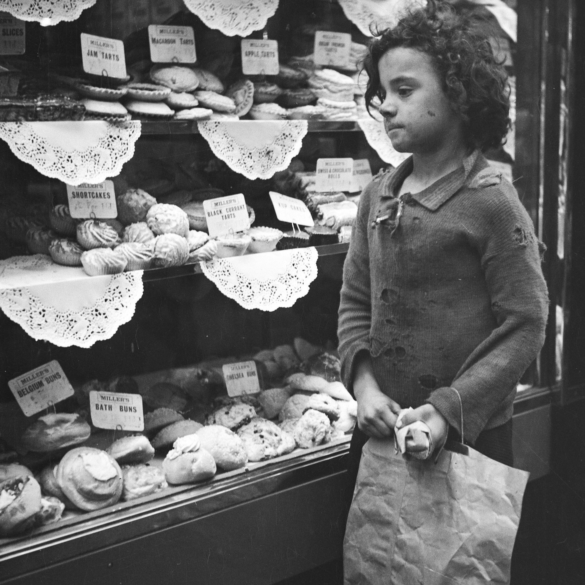 Die Schwarz-Weiß-Aufnahme von Edith Tudor-Hart zeigt ein kleines Mädchen in verschlissener Kleidung, das 1935 in London vor dem Schaufenster einer Bäckerei steht.