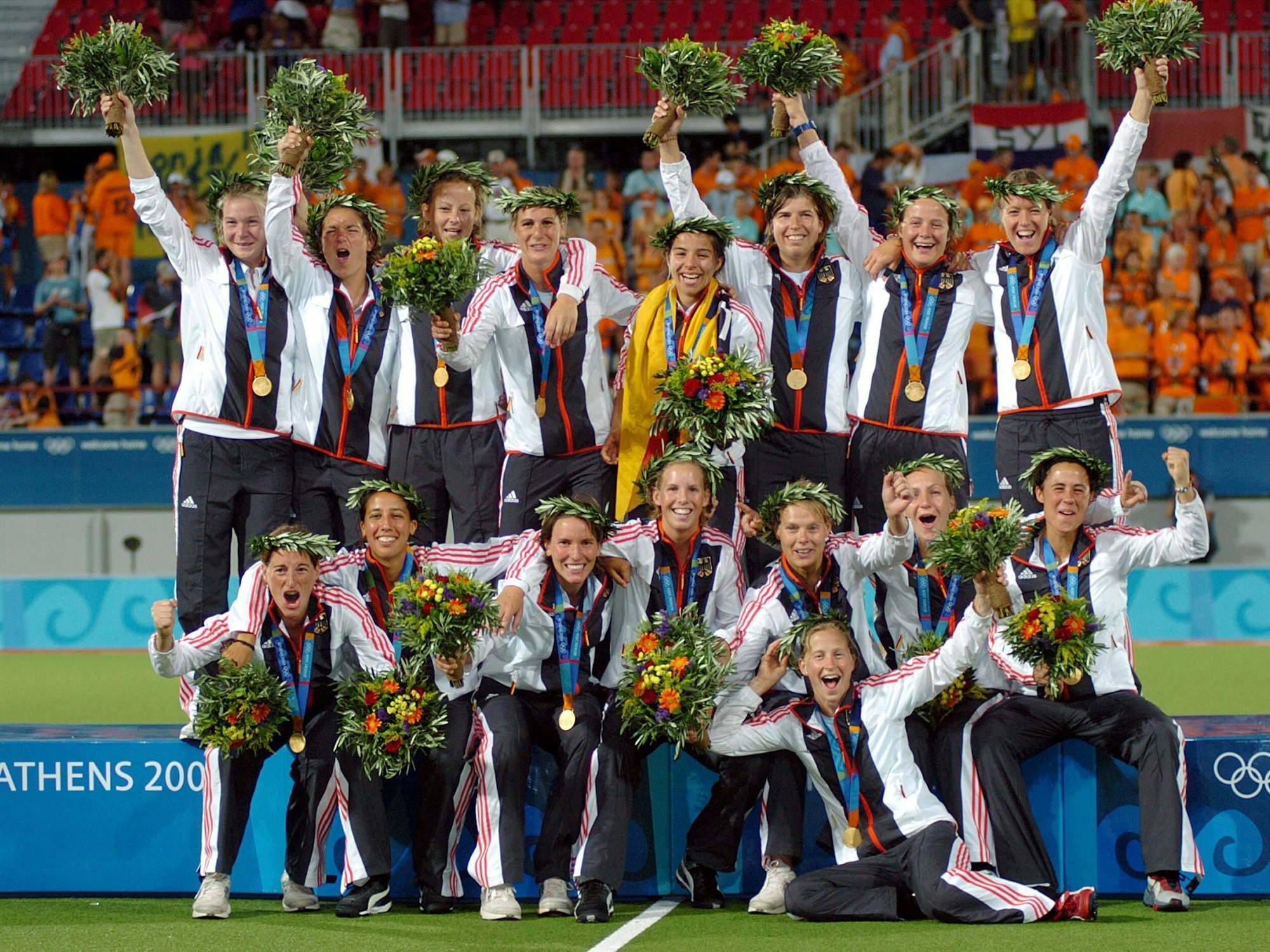 Das deutsche Hockey-Team der Damen feiert den Gewinn der Goldmedaille, mit Blumensträußen in den Händen und traditionellen Lorbeerkränzen auf den Köpfen.