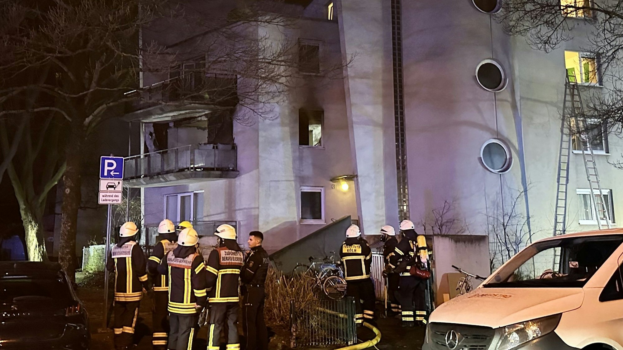 Einsatzkräfte der Kölner Feuerwehr stehen vor einem Haus in der Tiefentalstraße. Im Hintergrund ist eine ausgebrannte Wohnung zu sehen.