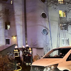 Einsatzkräfte der Kölner Feuerwehr stehen vor einem Haus in der Tiefentalstraße. Im Hintergrund ist eine ausgebrannte Wohnung zu sehen.