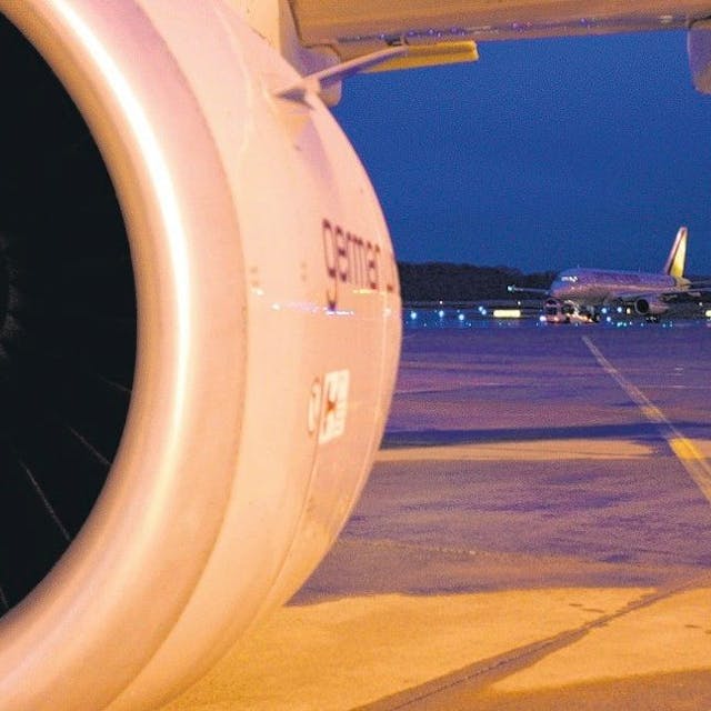 Das Bild zeigt die Turbine eines Flugzeuges und dahinter Vorfeld sowie Start- und Landebahn mit einem weiteren Flugzeug.