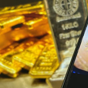 Auf einem Bildschirm sind die Edelmetalle Gold und Silber zu sehen, davor ist ein Smartphone-Bildschirm zu sehen, auf dem das Logo der Kryptowährung Bitcoin zu sehen ist.&nbsp;