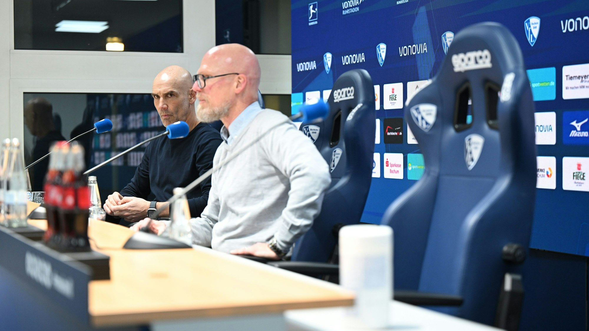 Bochums Trainer Thomas Letsch (l) und Bochums Pressesprecher Jens Fricke sitzen in der Pressekonferenz nach dem Spiel, der rechte Platz von Münchens Cheftrainer Tuchel ist leer.