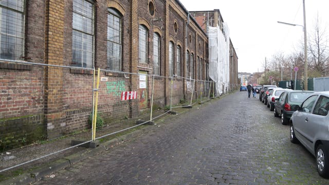Man sieht einen Bauzaun vor einem heruntergekommenen ehemaligen Industriegebäude.