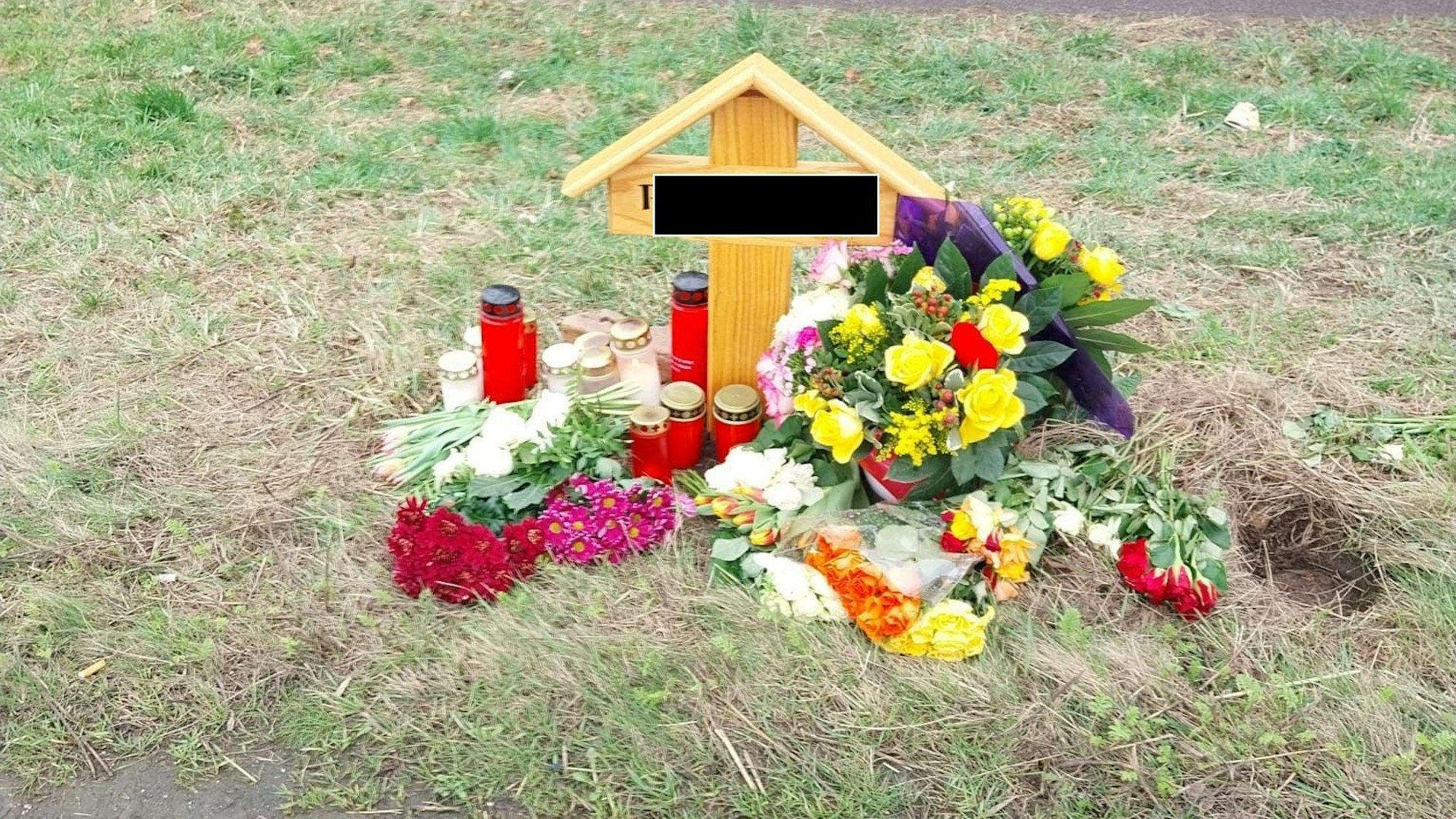 Ein Holzkreuz steht an einer Straße, an dem Kreuz wurden mehrere Blumensträuße niedergelegt und Kerzen aufgestellt.