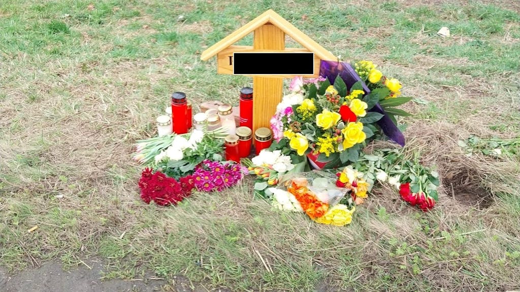 Ein Holzkreuz steht an einer Straße, an dem Kreuz wurden mehrere Blumensträuße niedergelegt und Kerzen aufgestellt.&nbsp;