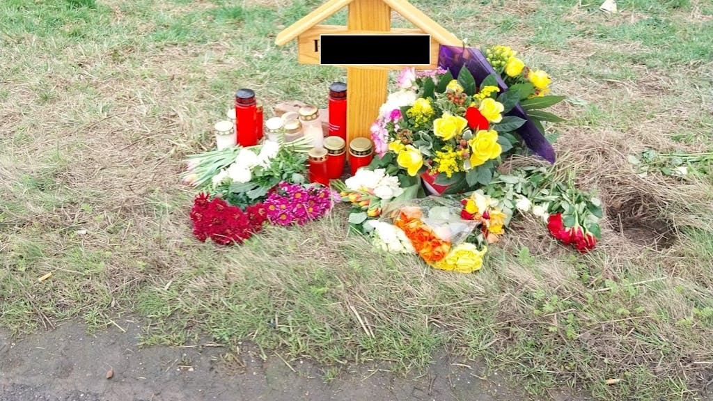Ein Holzkreuz steht an einer Straße, an dem Kreuz wurden mehrere Blumensträuße niedergelegt und Kerzen aufgestellt.&nbsp;