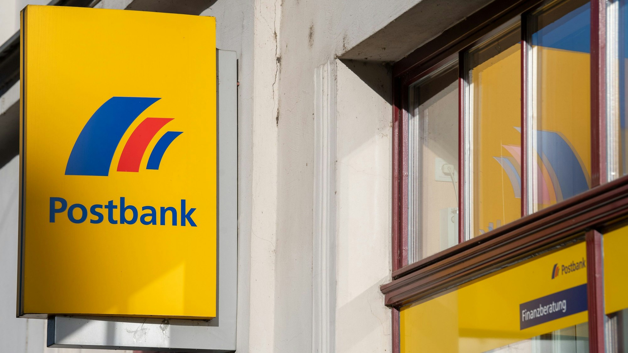 Ein Schild mit dem Logo der Postbank ist an der Fassade einer Bankfiliale angebracht.