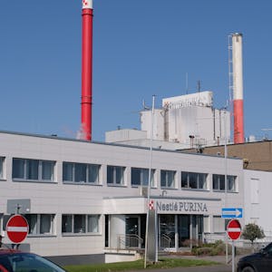 Das Bild zeigt den Eingang zum Verwaltungstrakt der Fabrik, im Hintergrund zwei Schornsteine.