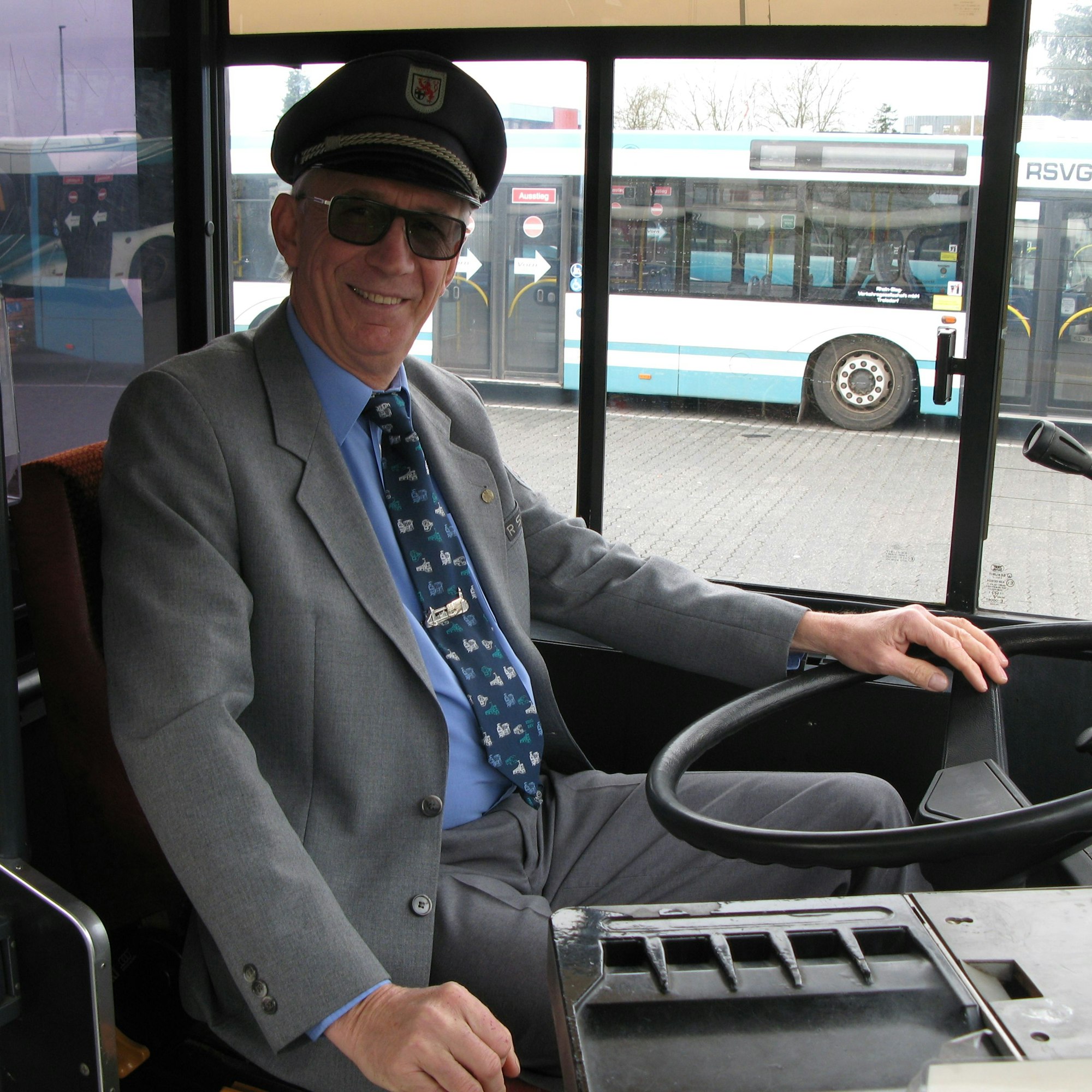 Verkehrsmeister Hans Willi Trost sitzt am Steuer des Oldtimers, er trägt seine alte Busfahrer-Uniform mit grauer Hose, grauem Jackett, blauem Hemd, Krawatte und Schirmmütze.