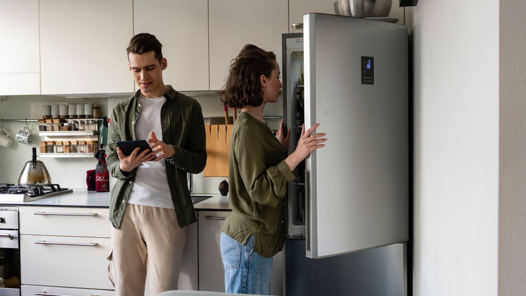 Auf dem Foto sieht man eine Frau einen Kühlschrank öffnen, neben ihr steht ein Mann, welcher auf sein Tablet schaut.