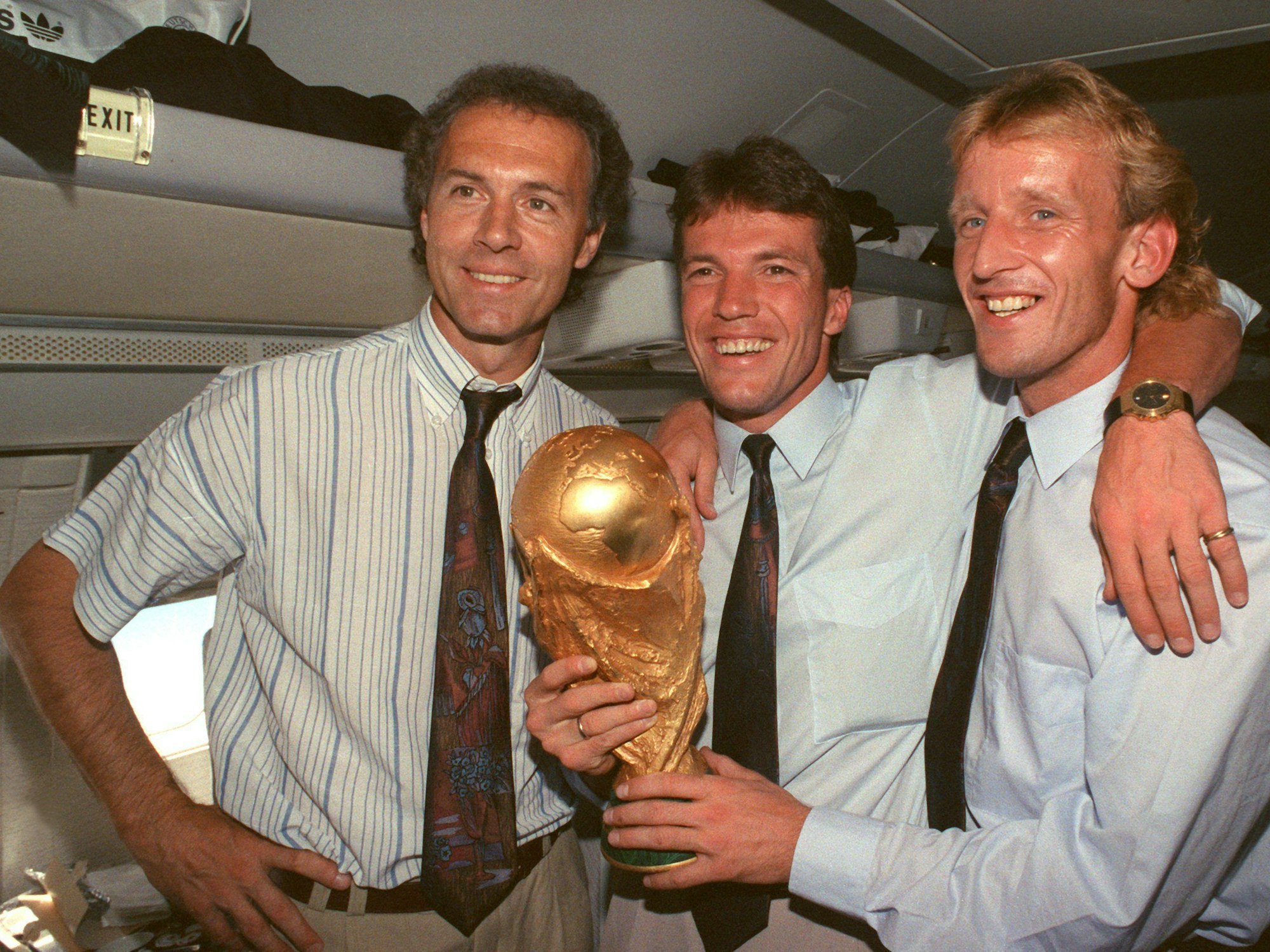 Franz Beckenbauer, Lothar Matthäus und Andreas Brehme halten auf dem Flug von Rom nach Frankfurt den WM-Pokal in der Hand.